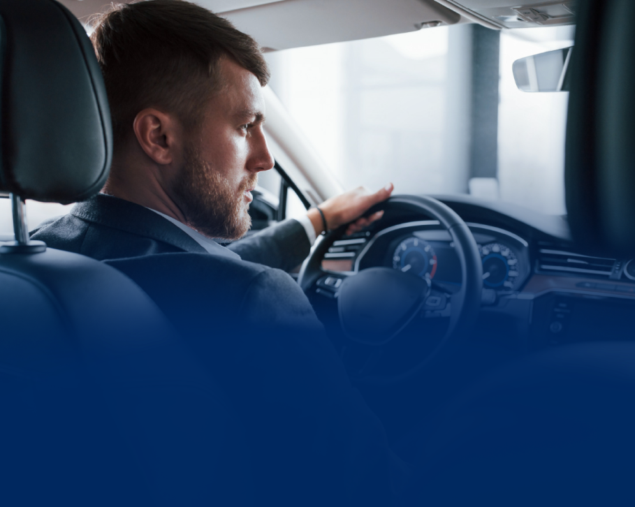 Homem dirigindo um carro, segurando o volante, vista de perfil lateral, traje formal.