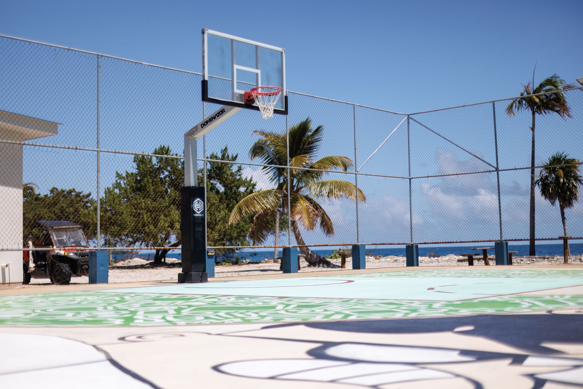 Outdoor basketball court by the ocean with a hoop, palm trees, and blue sky