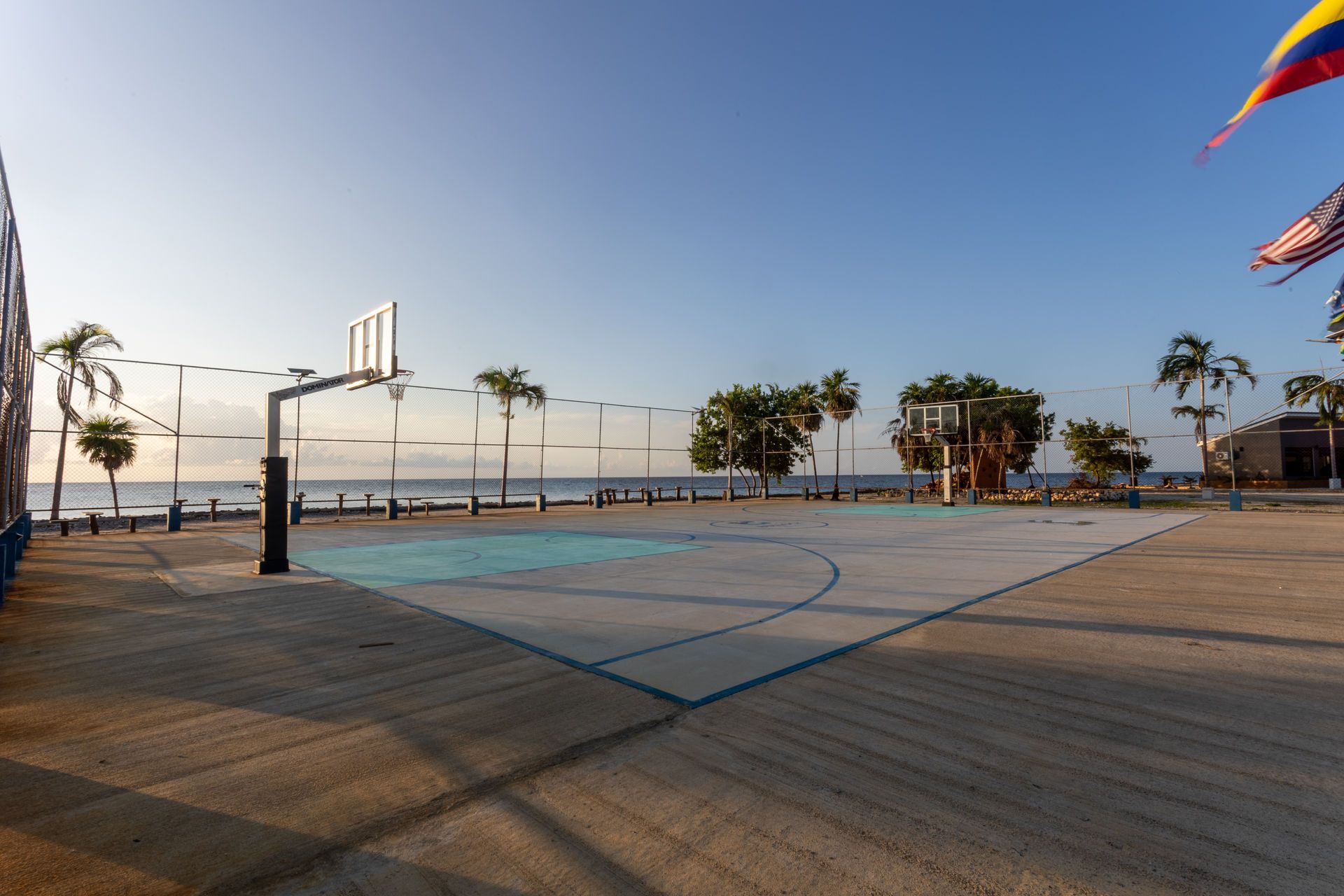 Basketball court by the ocean, with palm trees and flags in the background. Blue and light-colored court.