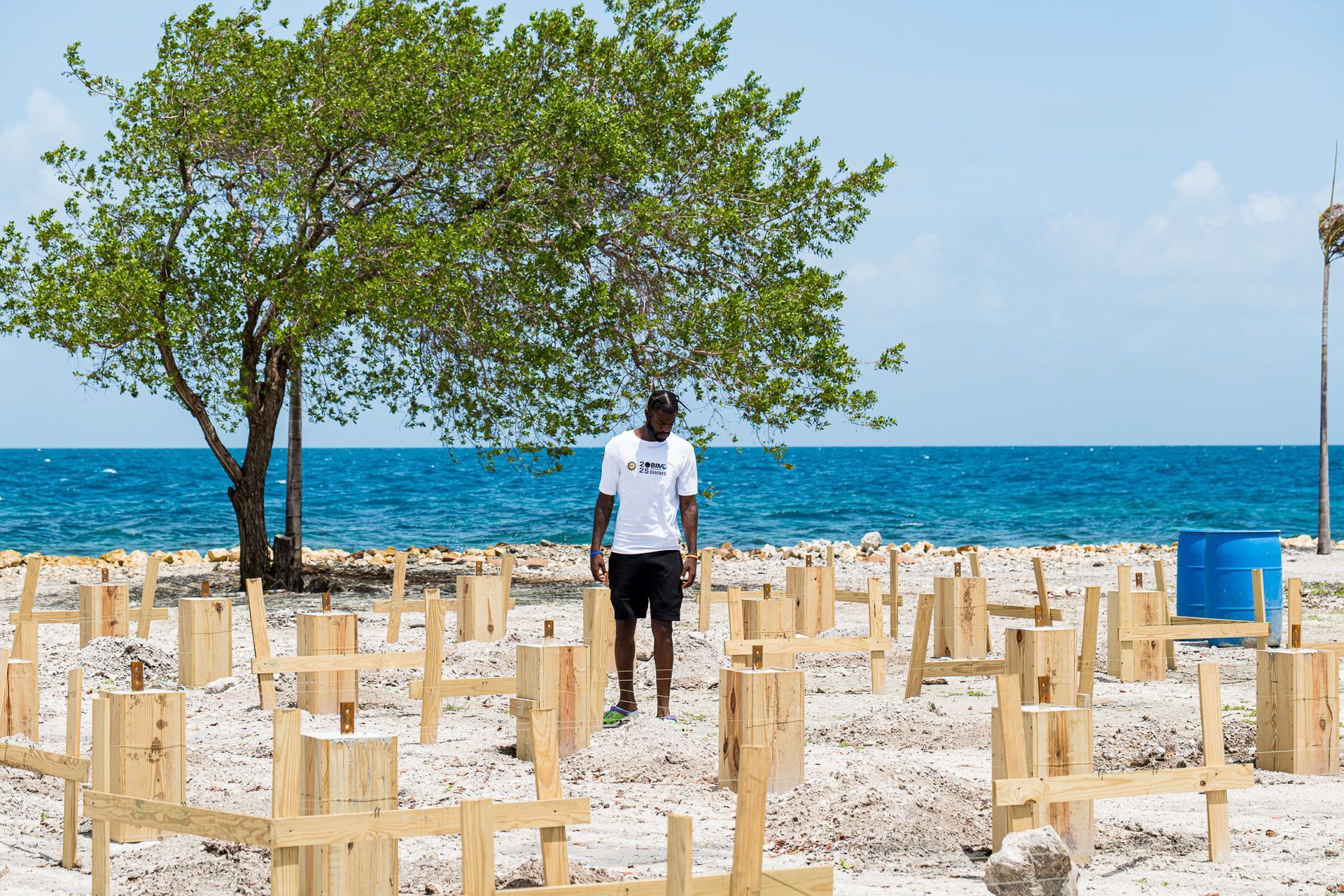 Man standing on beach among wooden structures, tree, ocean background. Bright sunny day.