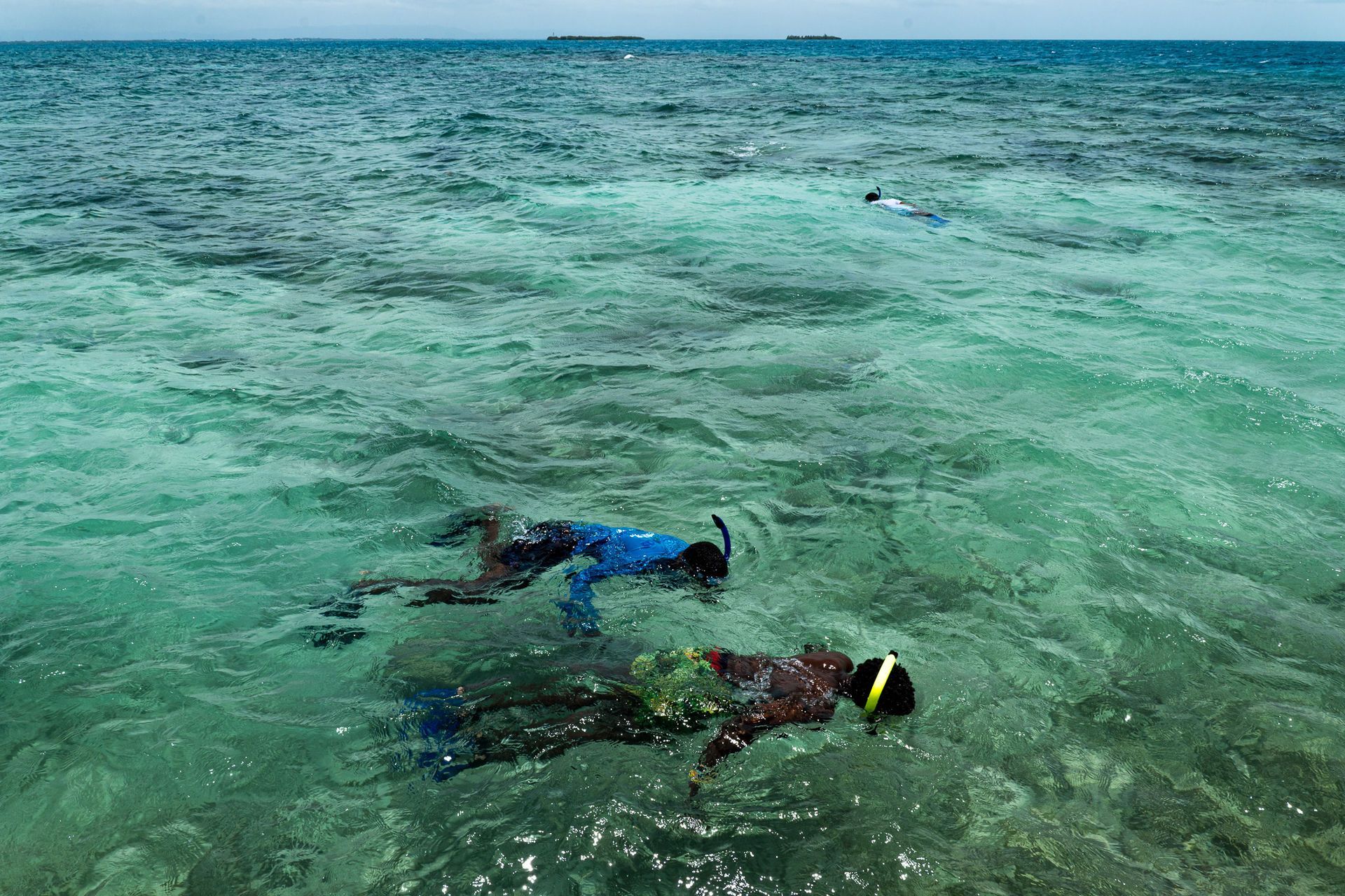 Two people snorkeling in clear, turquoise water. One wears a blue shirt, the other a green shirt.