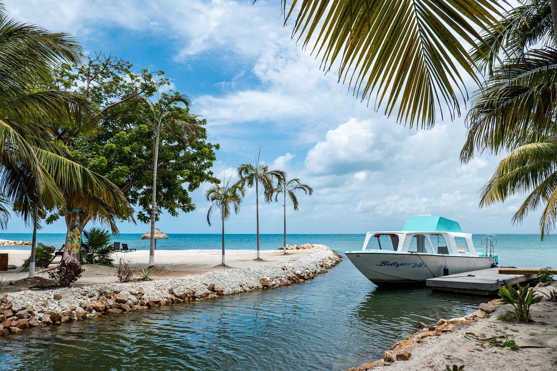 Boat docked in a canal with palm trees, sandy shore, and ocean visible under a blue sky.