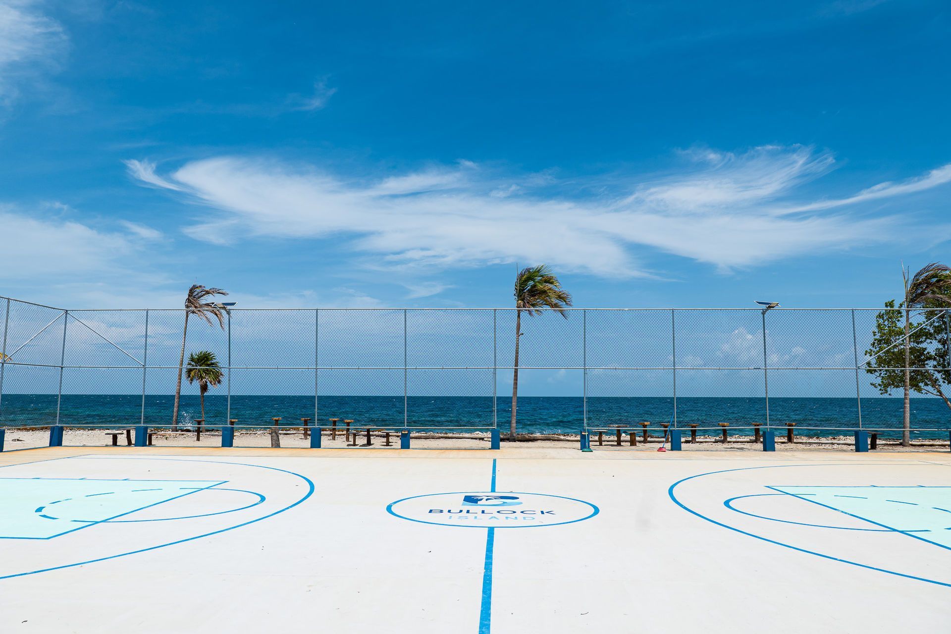 Basketball court by the ocean, blue and white painted lines, net and fence, palm trees, sunny sky.
