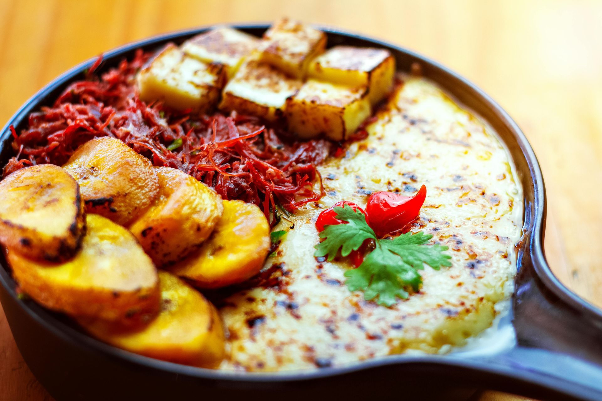 A skillet with a dish of fried plantains, shredded meat, cheese, and fried cheese cubes, with a red pepper garnish.