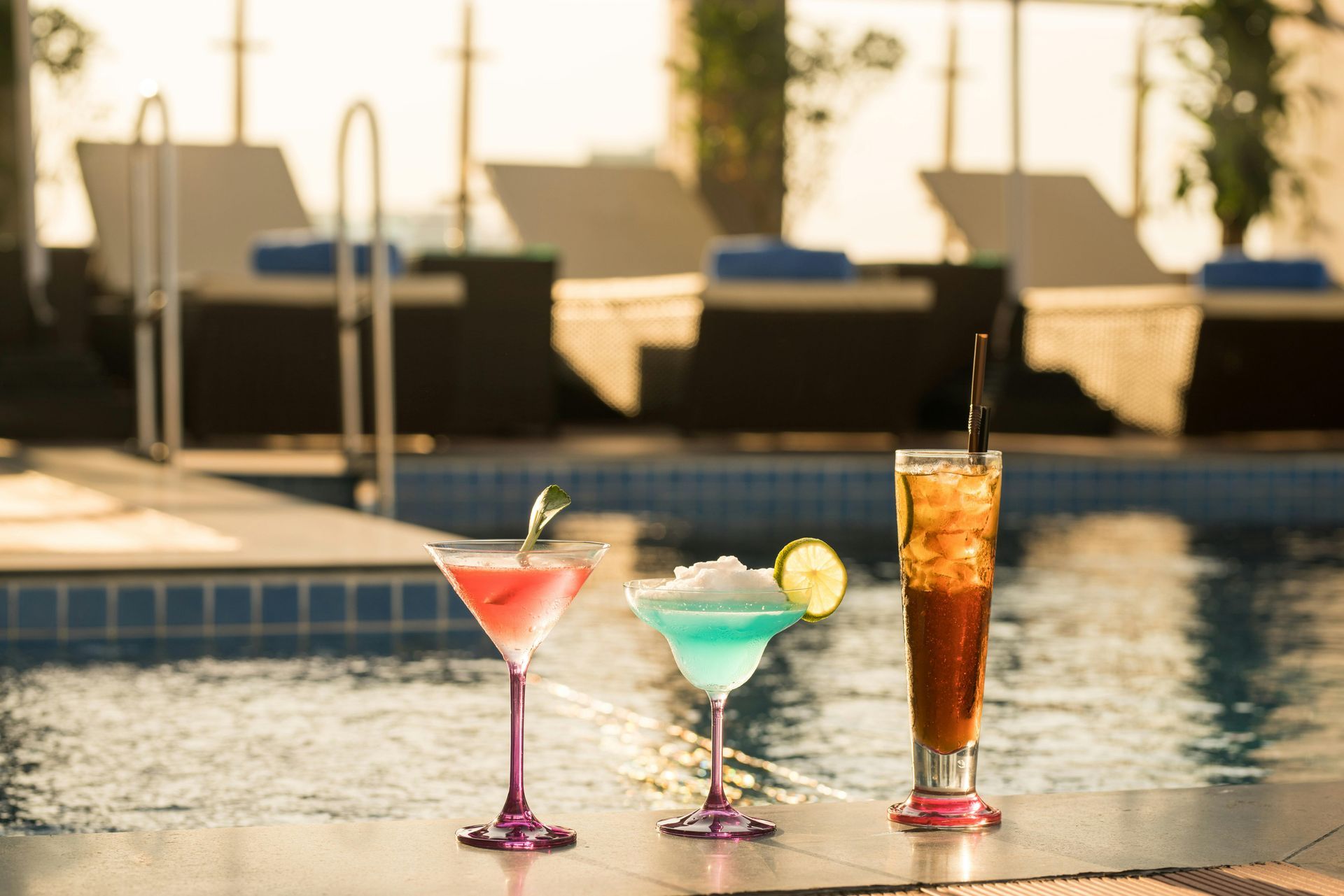 Three colorful cocktails by a pool with lounge chairs in the background.