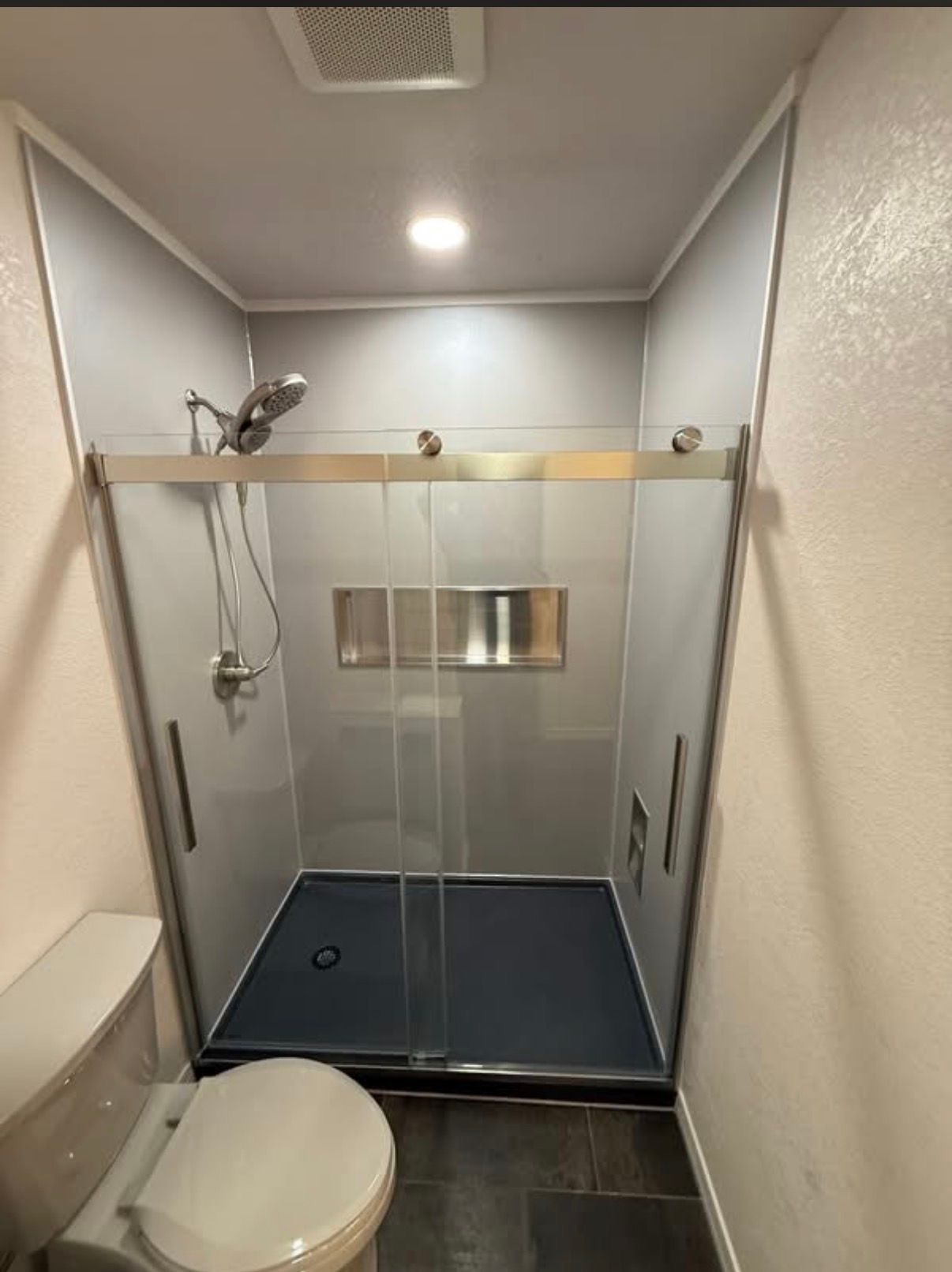A walk-in shower with light grey walls, a dark base, silver trim, sliding glass doors, and a built-in recessed wall niche.