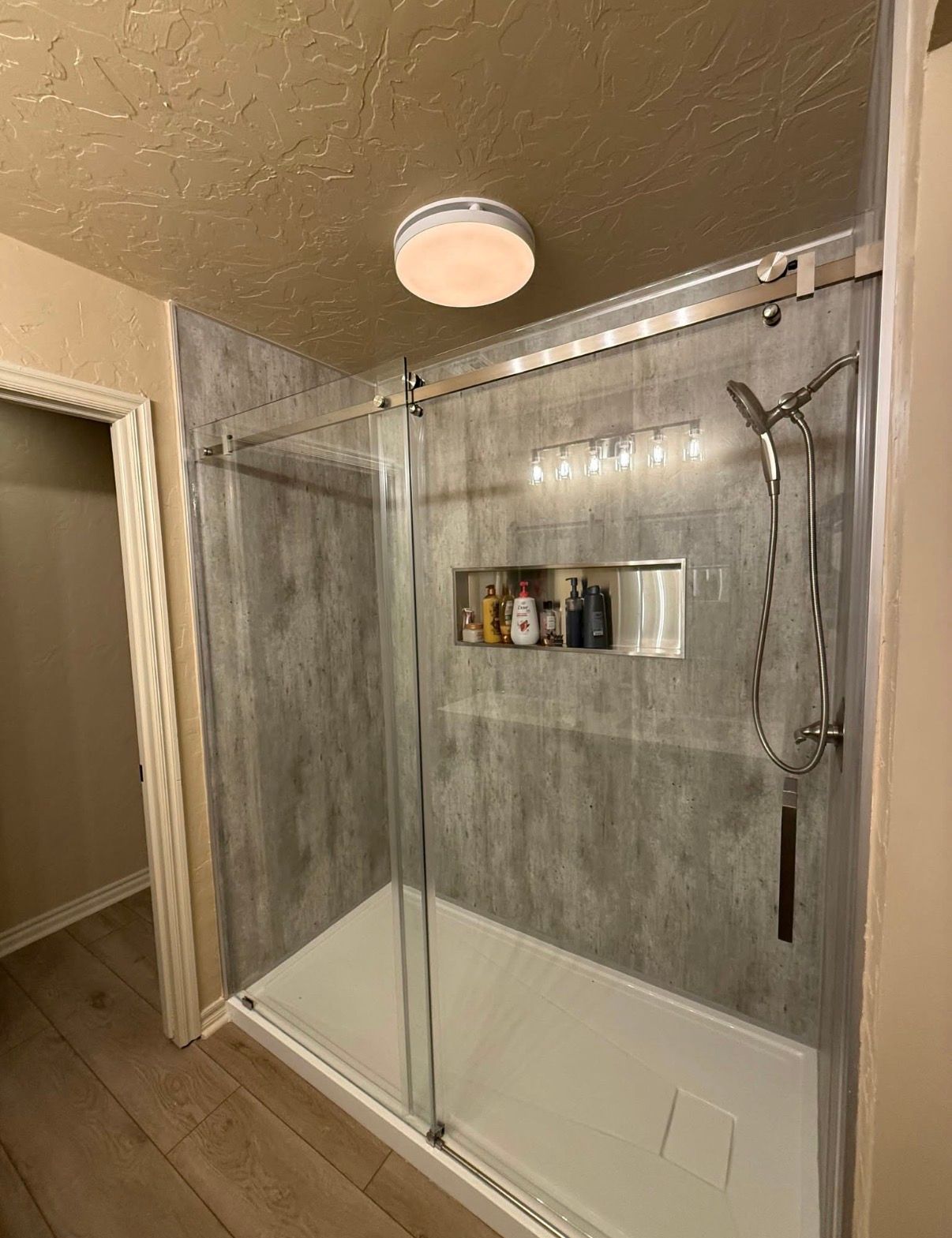 A walk-in shower with gray marbled walls, sliding glass doors, a handheld showerhead, and a recessed storage niche.