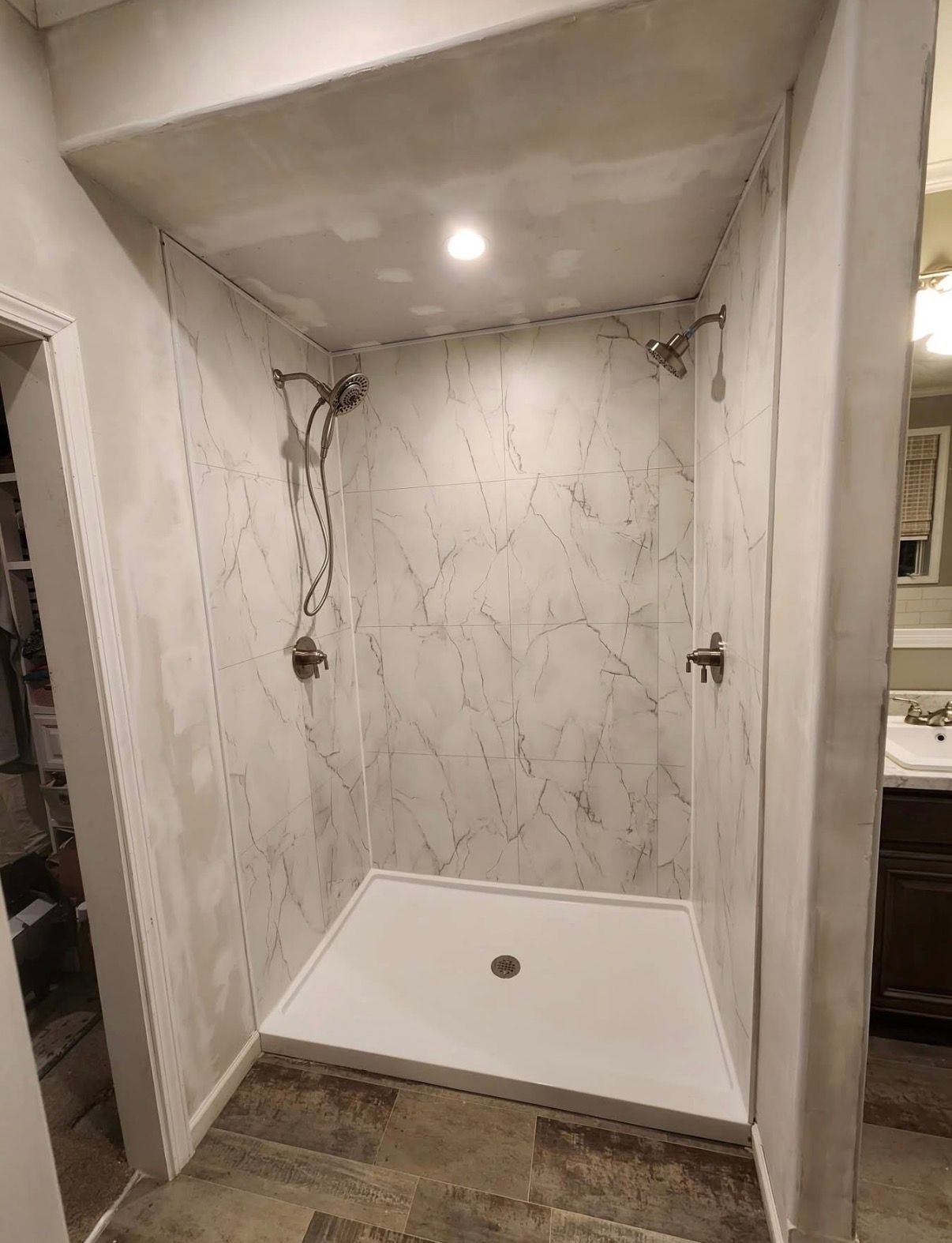 A walk-in shower with marble-patterned walls, two chrome showerheads, a white base, and a recessed ceiling light.