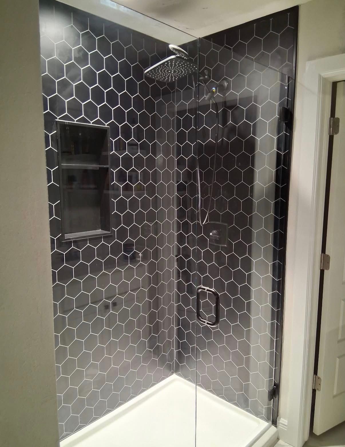 A walk-in shower with black hexagonal tiles, a glass door, a built-in shelf, and a square chrome shower head.