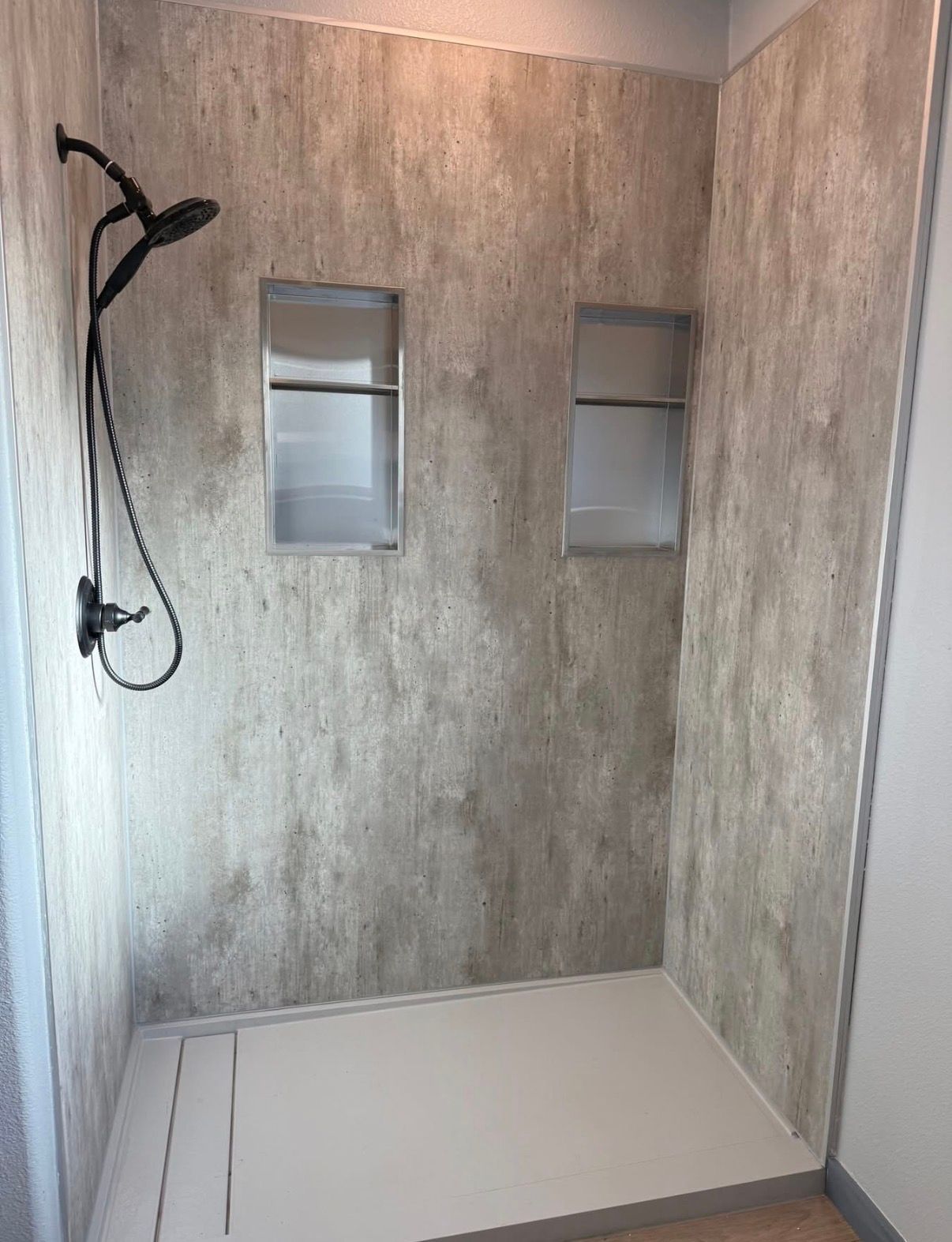 A walk-in shower with light brown textured stone-look walls, a white floor, two recessed shelves, and a black showerhead.