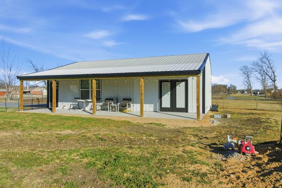 A white barndominium with a covered front porch and metal roof set in a grassy yard under a blue sky.
