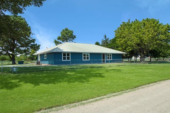 A blue, one-story house with a light-colored roof, surrounded by a green lawn, trees, and a chain-link fence under a blue sky.