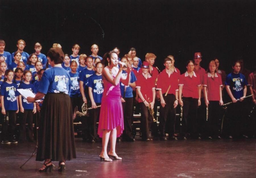 A woman in a pink dress sings into a microphone in front of a choir