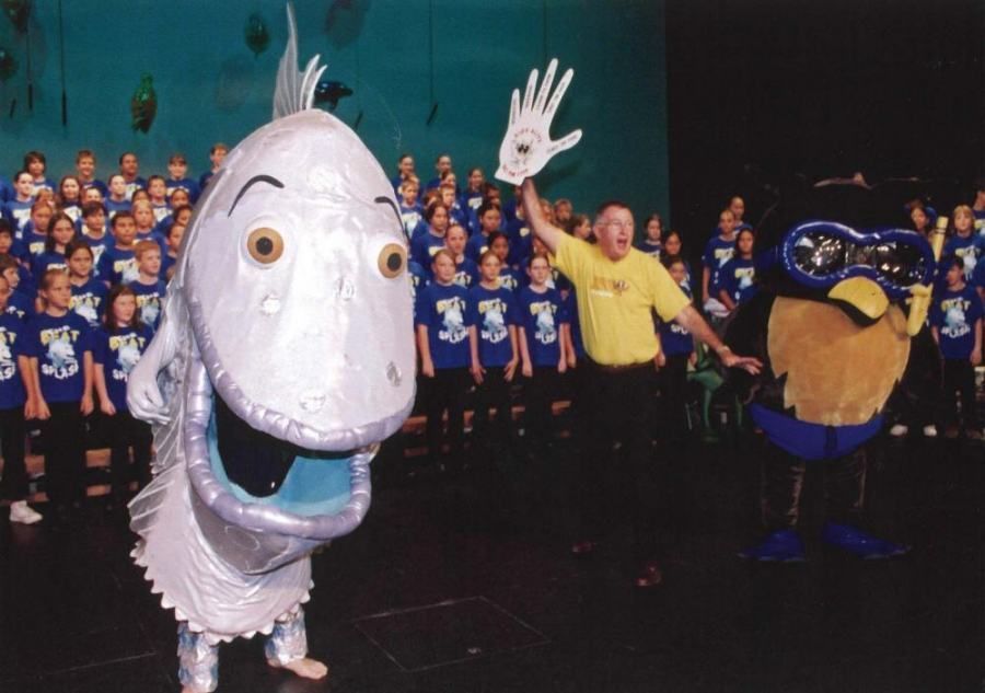 A man in a yellow shirt is standing next to a fish mascot
