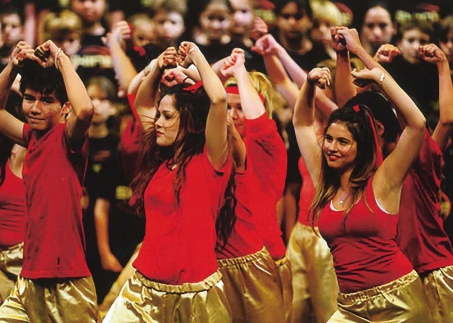 A group of people in red shirts and gold pants are dancing in front of a crowd