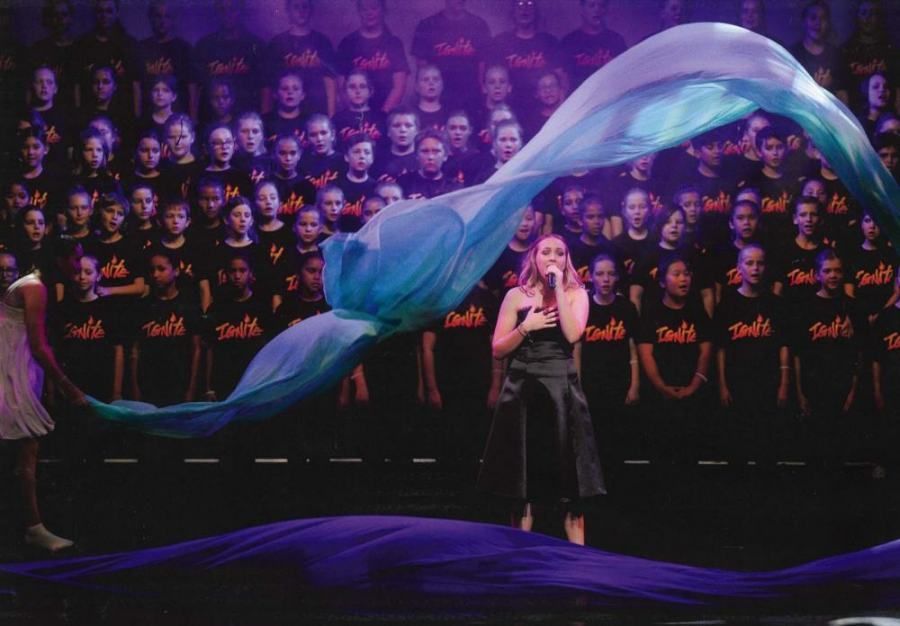 A woman is singing into a microphone in front of a large choir.