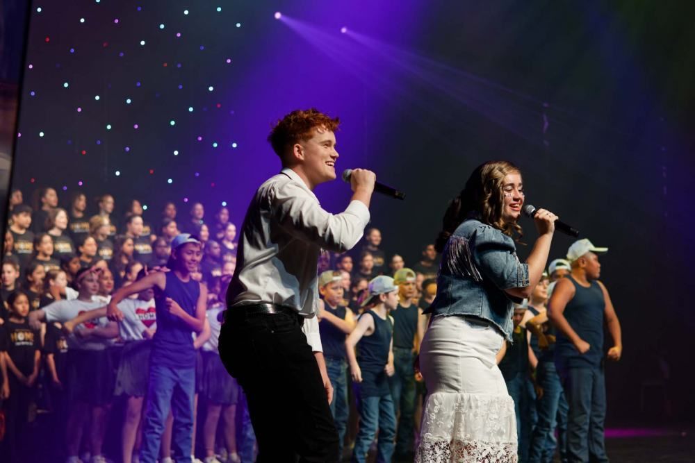 A boy and a girl are singing into microphones on a stage in front of a choir.