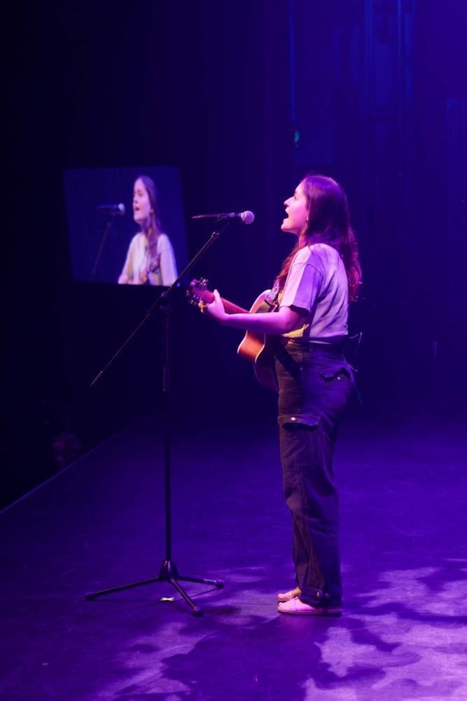 A woman is playing a guitar and singing into a microphone on a stage.
