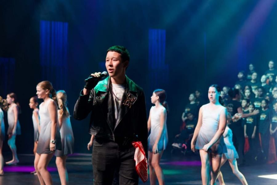 A man is singing into a microphone on a stage in front of a group of dancers.