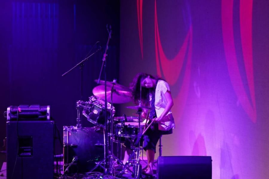 A woman is playing drums on a stage with purple lights behind her.