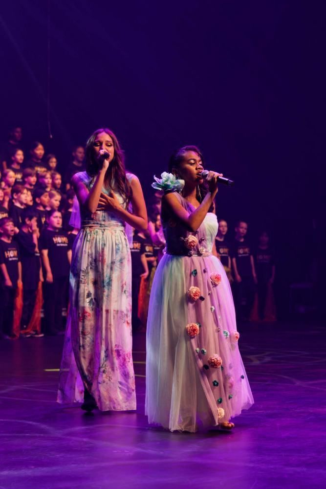 Two women are singing into microphones on a stage in front of a choir.