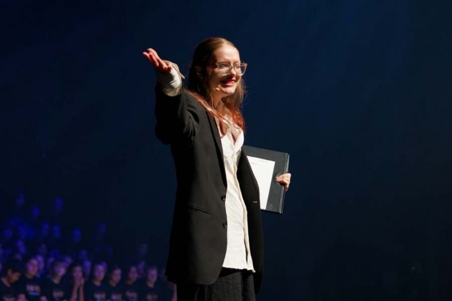 A woman in a suit is standing on a stage holding a clipboard.