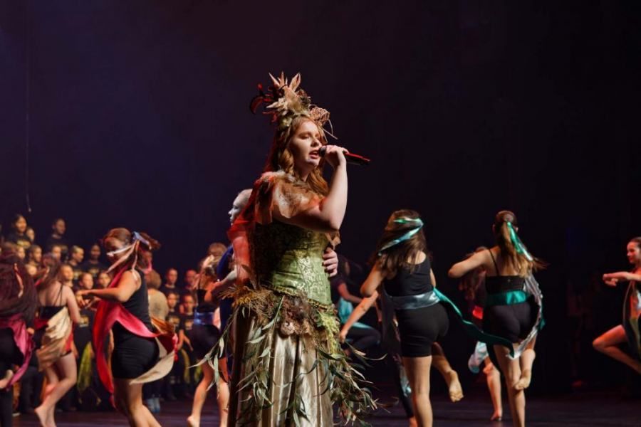 A woman is singing into a microphone while a group of dancers are dancing in the background.