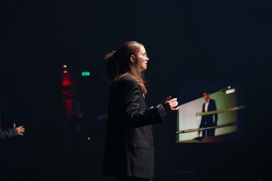 A woman in a black suit is giving a speech on a stage.