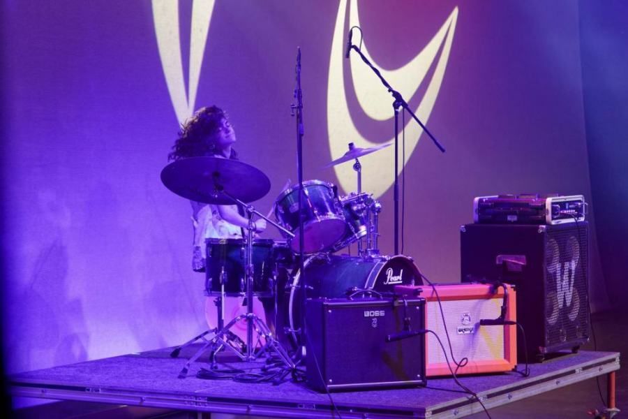 A man is playing drums on a stage in front of a purple background.