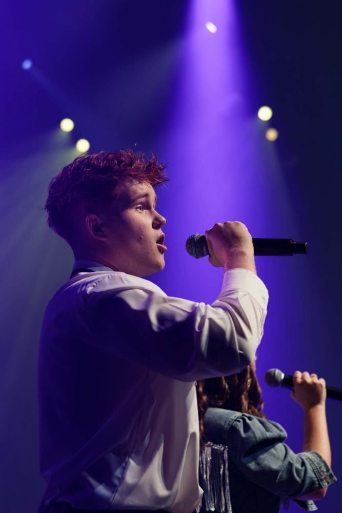 A man singing into a microphone on a stage with purple lights behind him