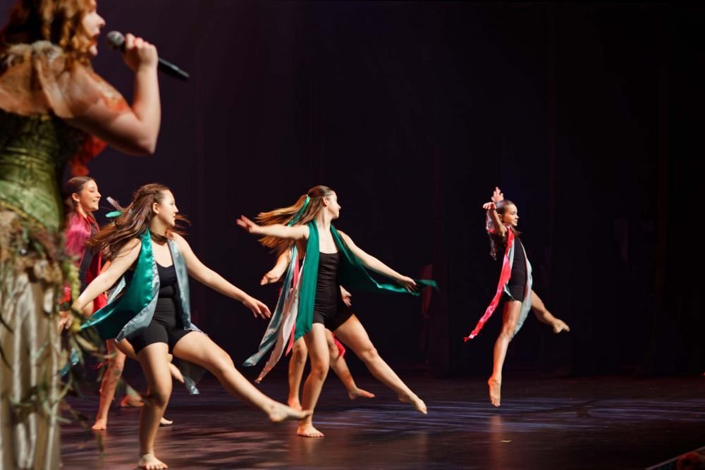 A group of young girls are dancing on a stage while a woman sings into a microphone.