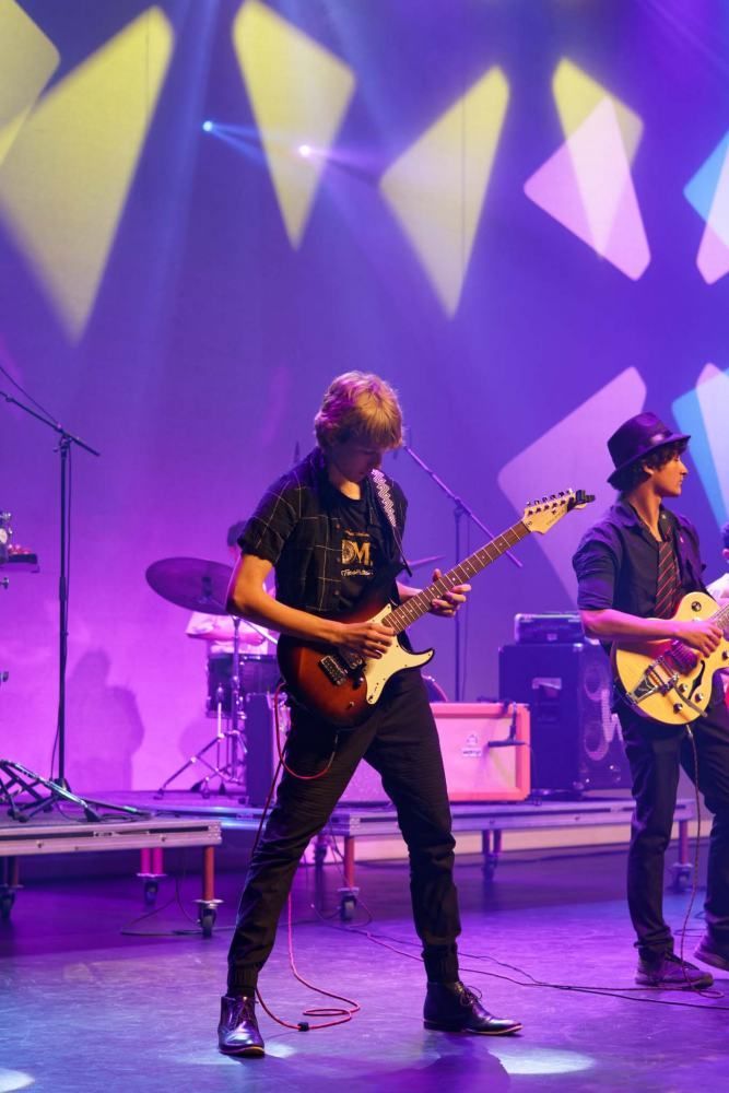 Two men are playing guitars on a stage with purple lights behind them.