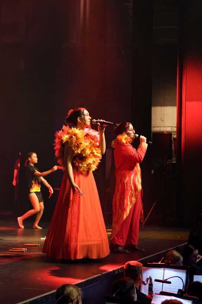 Two women in red dresses are singing into microphones on a stage.