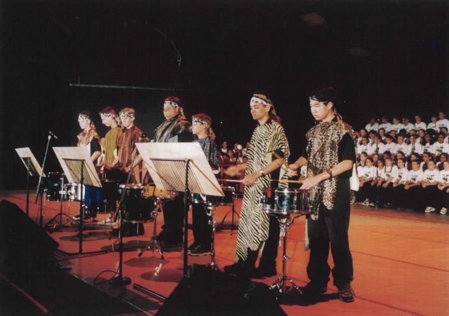 A group of people playing drums on a stage with a choir behind them