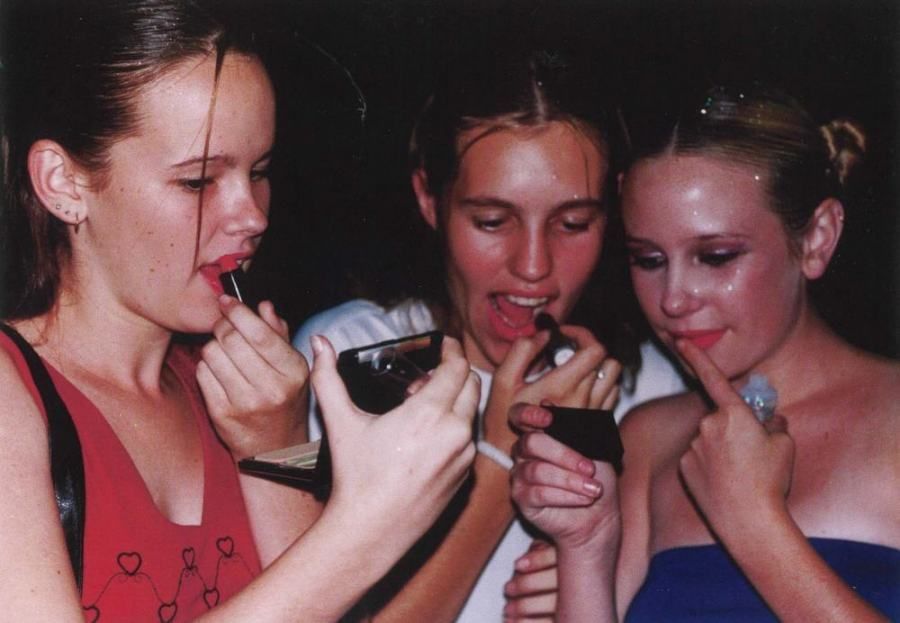 Three girls are applying lipstick and looking at a cell phone