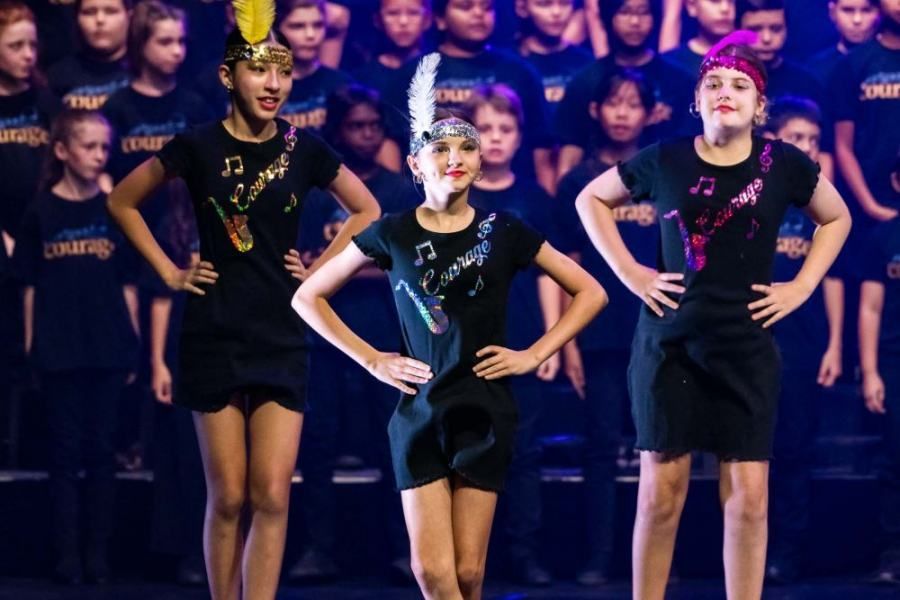 Three young girls are standing on a stage in front of a choir.