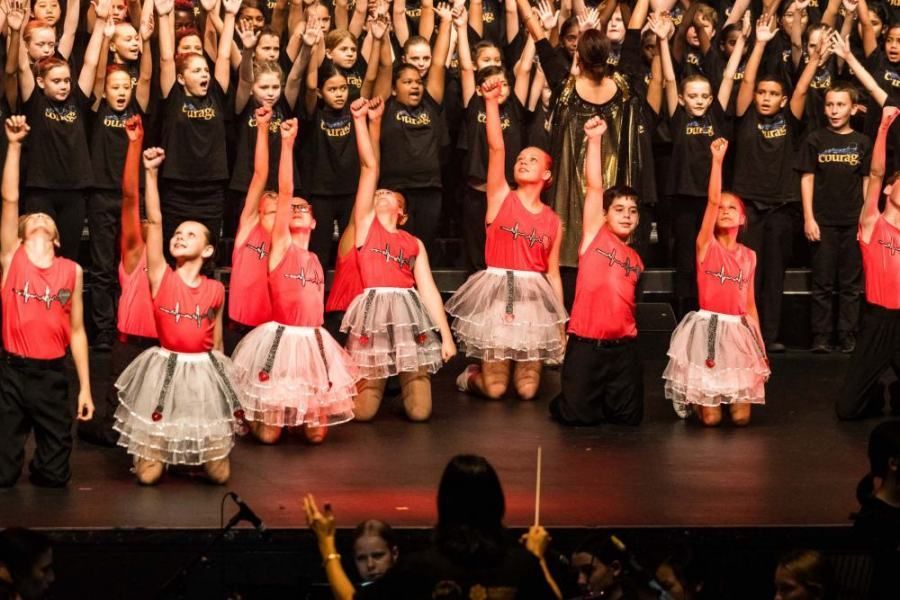 A large group of children are standing on a stage with their hands in the air.