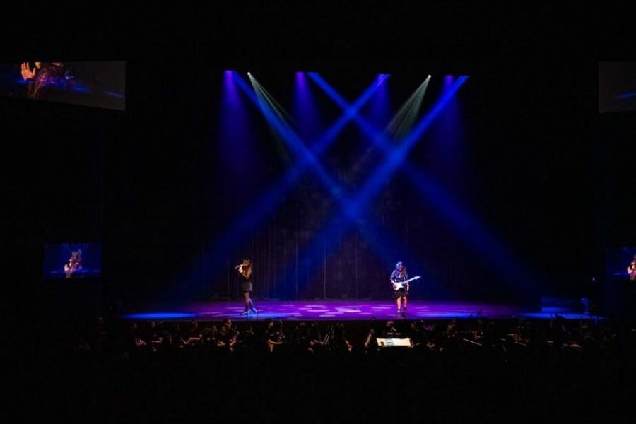 Two people are playing guitars on a stage in front of a crowd.