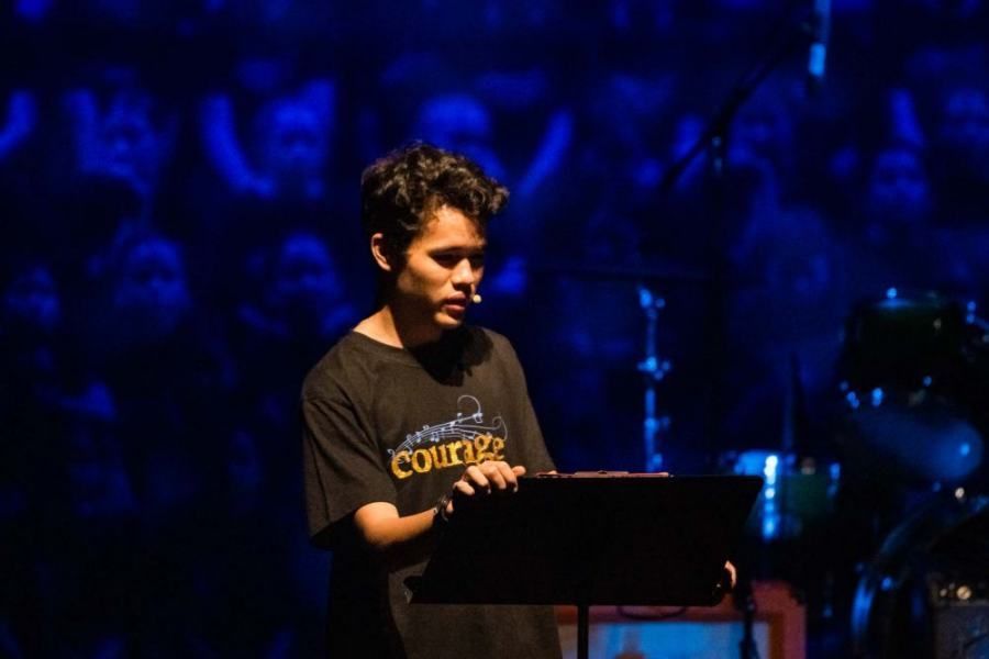 A man wearing a black shirt that says course is holding a laptop on a stage.