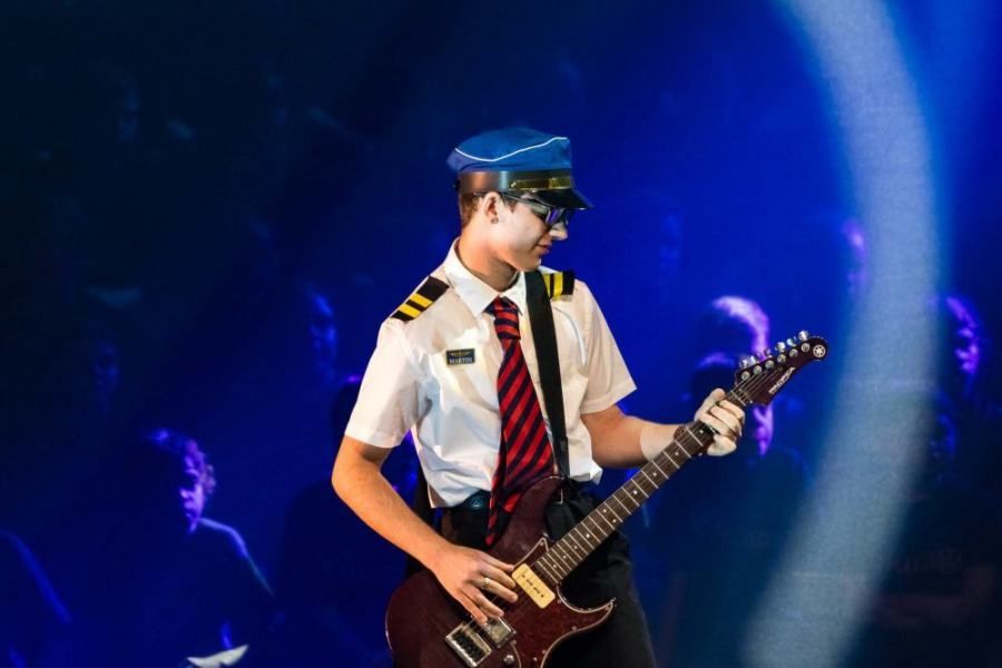 A man in an airplane uniform is playing a guitar on a stage.