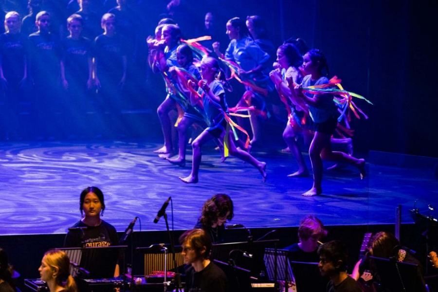 A group of children are dancing on a stage in front of an orchestra.