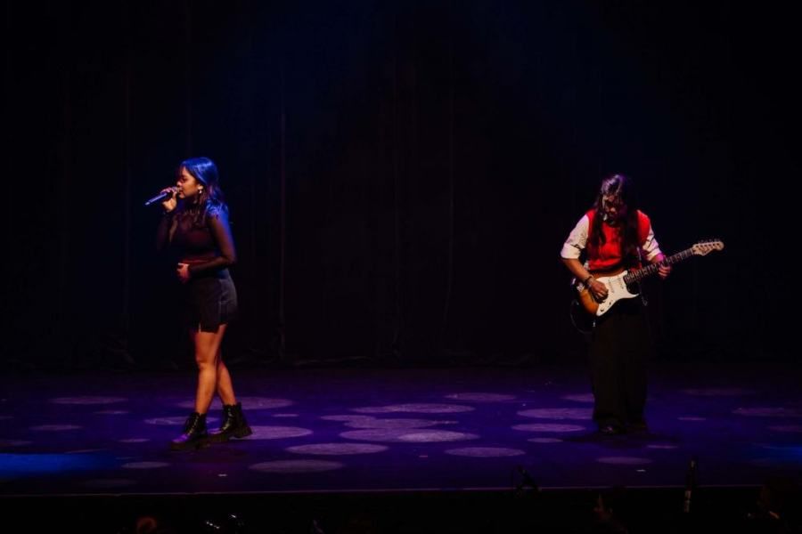 A woman is singing into a microphone next to a man playing a guitar on a stage.