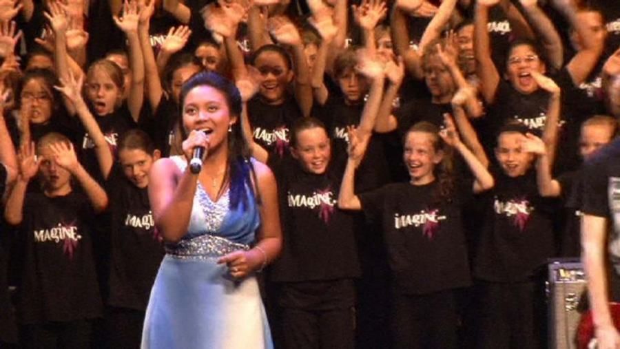 A woman singing into a microphone in front of a crowd of children wearing black shirts that say imagine