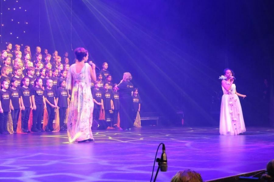 Two women singing on a stage with a choir behind them