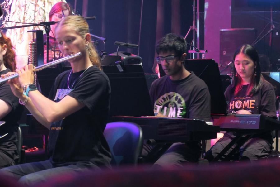 A woman is playing a flute in an orchestra while a man is playing a keyboard.
