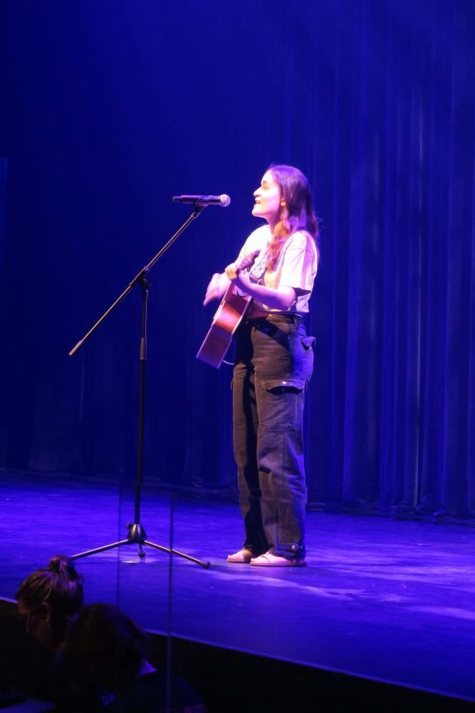 A woman is singing into a microphone while holding a guitar on a stage.