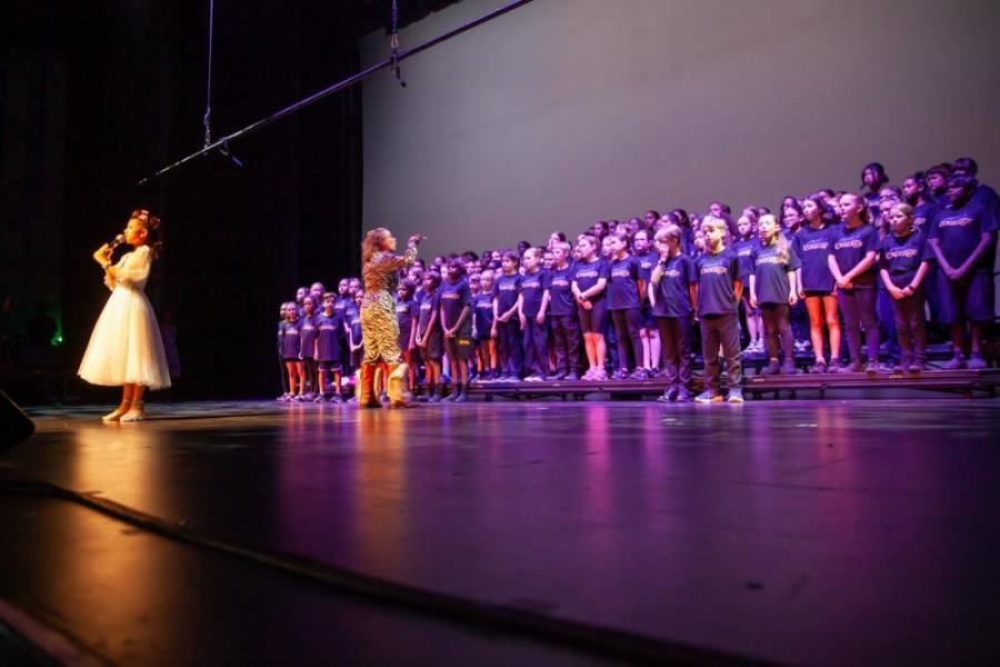 A woman is singing into a microphone in front of a large group of children on a stage.
