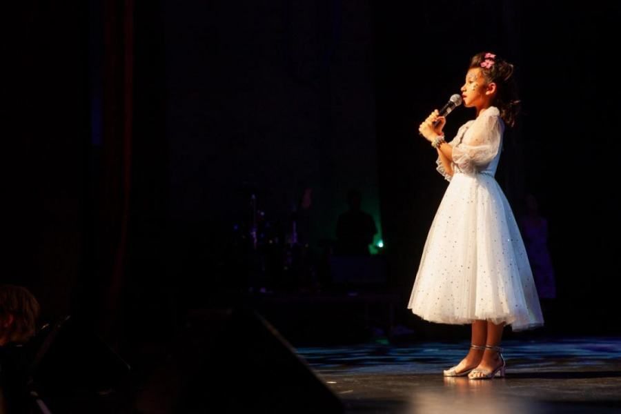A little girl in a white dress is singing into a microphone on a stage.