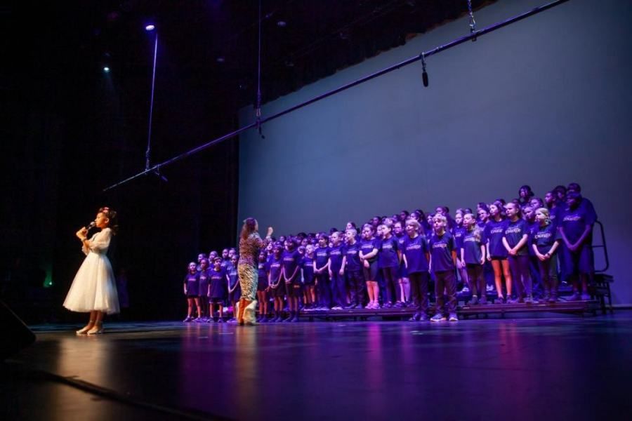 A woman is singing into a microphone on a stage in front of a large choir.