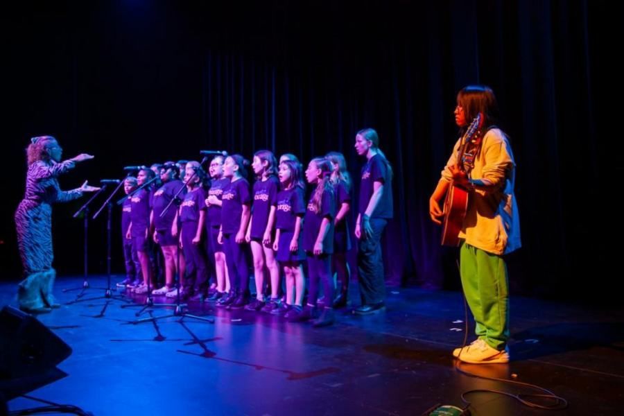 A group of children are singing on a stage with a man playing a guitar.